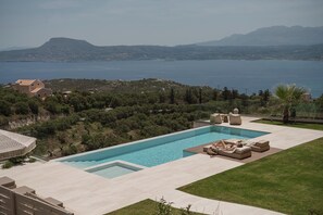 A swimming pool with a view of the sea and mountains, surrounded by a well-maintained lawn and outdoor furniture.
