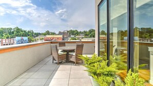 Una terraza en la azotea con una mesa redonda y sillas, una pared de vidrio y vistas a la ciudad.