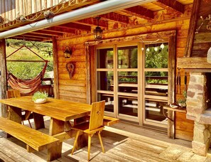 Una cabaña de madera con mesa y bancos, una hamaca y una chimenea de piedra.