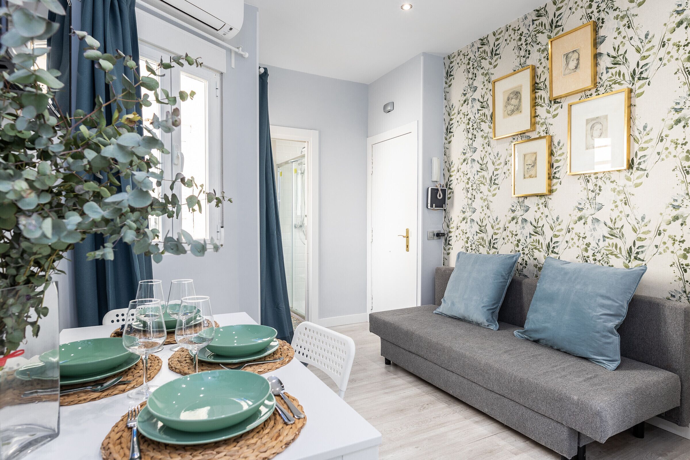 Un salon moderne avec un canapé gris, une table à manger dressée avec de la vaisselle verte et des verres, et des tableaux encadrés accrochés au mur.