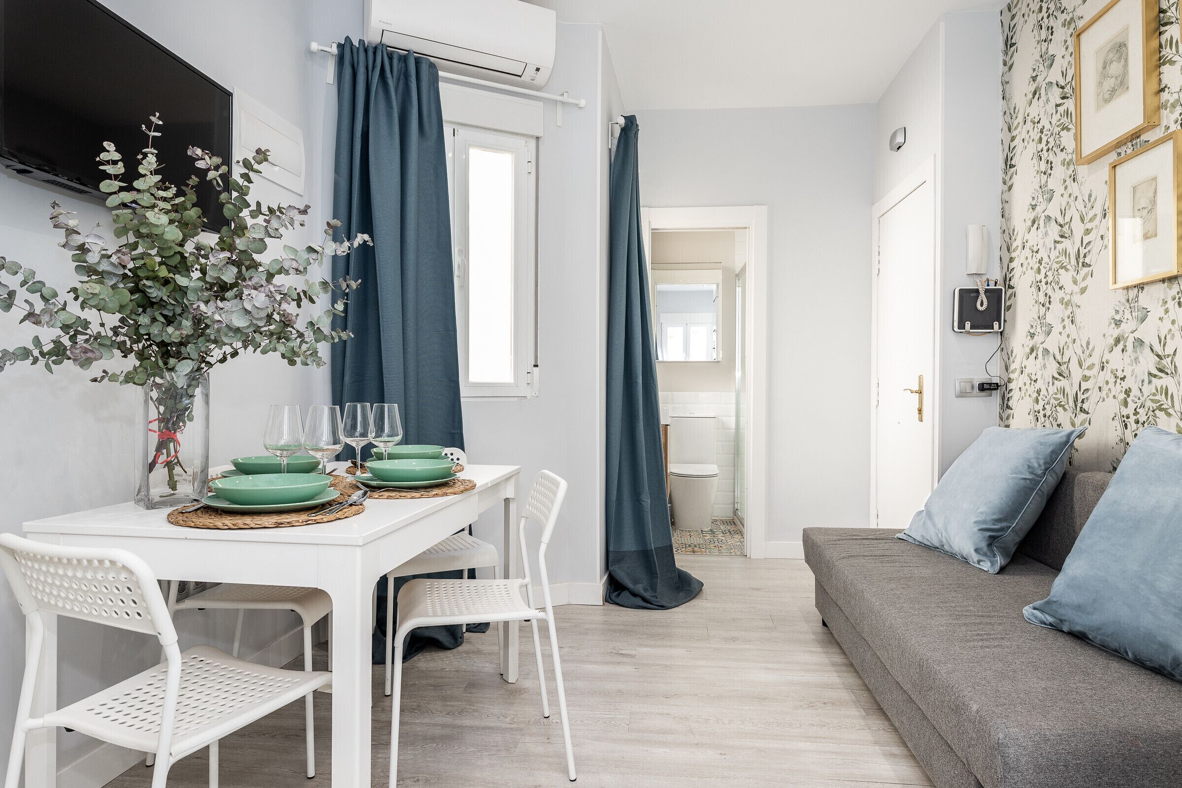 Un salon moderne avec un canapé gris, des chaises blanches, une table à manger avec de la vaisselle verte et des verres, et une grande fenêtre avec des rideaux.