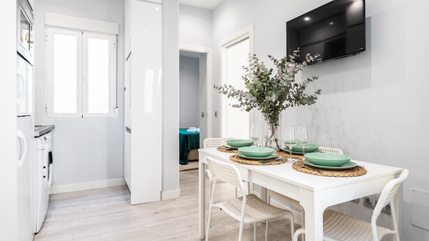 Une cuisine moderne avec des appareils électroménagers blancs, un coin repas avec une table et des chaises blanches, et un téléviseur à écran plat fixé au mur.