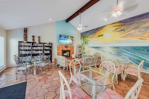 A living room with a fireplace, a large bookshelf, a dining area with chairs, and a mural of a sunset over the ocean.
