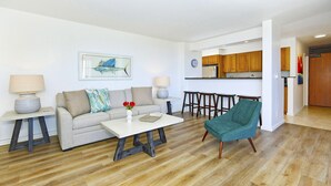 Ein modernes Wohnzimmer mit einer Couch, einem Couchtisch und einem petrolfarbenen Sessel. Im Hintergrund ist eine Küche mit Holzschränken und einem Kühlschrank erkennbar.
