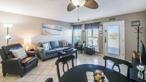 A living room with a sofa, armchairs, a coffee table, and a dining table. There is a ceiling fan, a painting, and a door with a sign. - Dog-Friendly Beachside Retreat | Patio, Pool & Hot Tub (Corpus Christi)