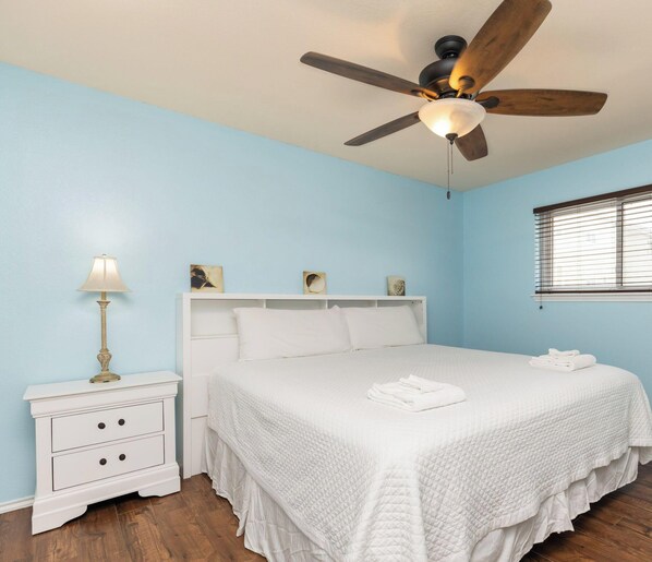 A bedroom with a bed, a nightstand, a lamp, a ceiling fan, and a window with blinds.