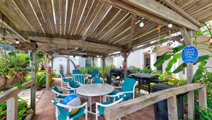 An outdoor patio area with wooden pergola, white plastic chairs, and a barbecue grill. - Steps to Beach & Pool | Easy Coastal Getaway (Corpus Christi)