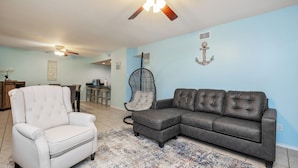 A living room with a white armchair, a dark leather sofa, a patterned rug, and a ceiling fan. - Steps to Beach & Pool | Easy Coastal Getaway (Corpus Christi)