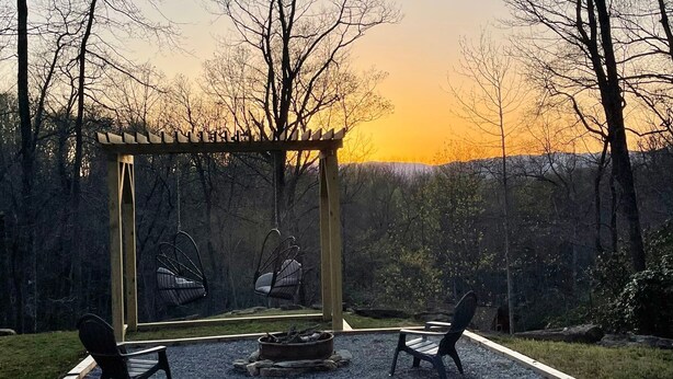 Un ensemble de jeux d’extérieur en bois, avec une structure en m étal, deux chaises et un foyer, installé dans un jardin offrant une vue sur le coucher du soleil.