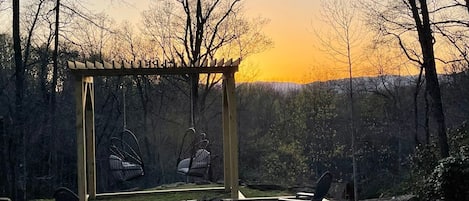 Un ensemble de jeux d’extérieur en bois, avec une structure en métal, deux chaises et un foyer, installé dans un jardin offrant une vue sur le coucher du soleil.