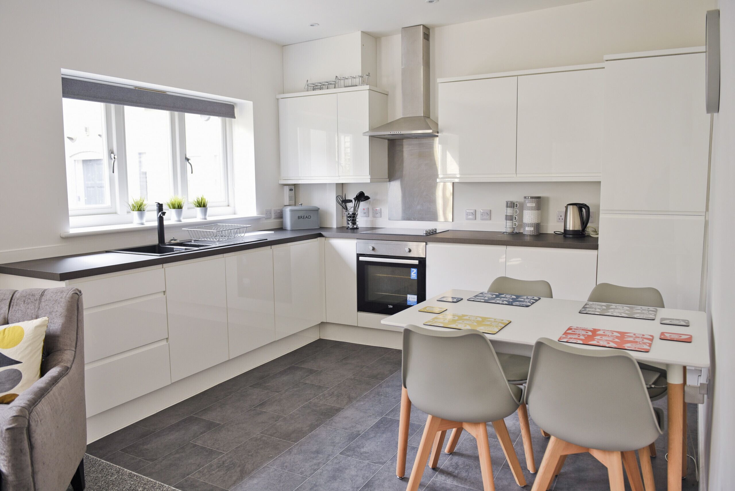 Een moderne keuken met witte kastjes, een ingebouwde oven, een rvs afzuigkap en een eethoek met een tafel en stoelen.