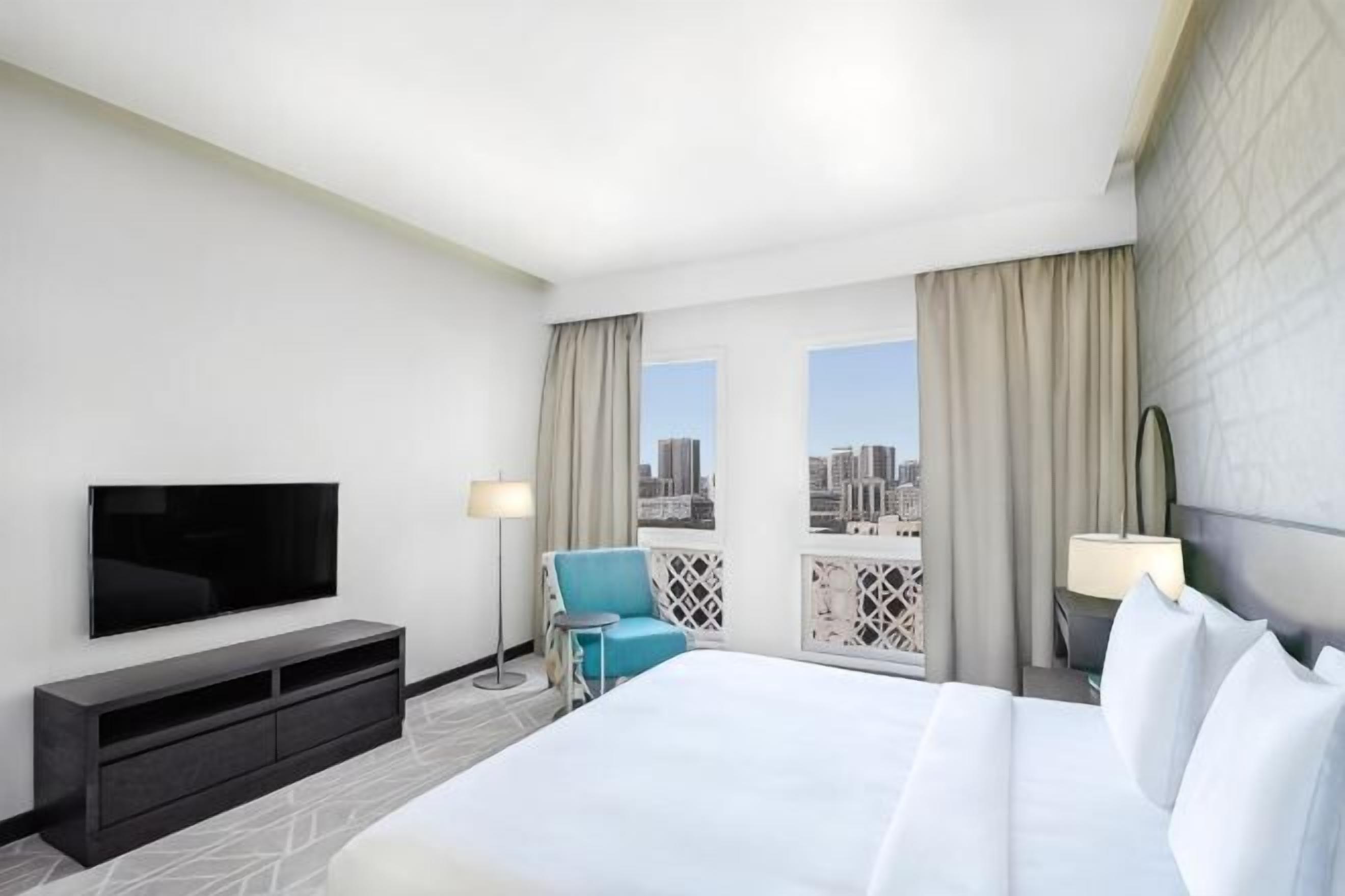 A hotel room with a large bed, a TV, a desk, and a balcony with a city view.
