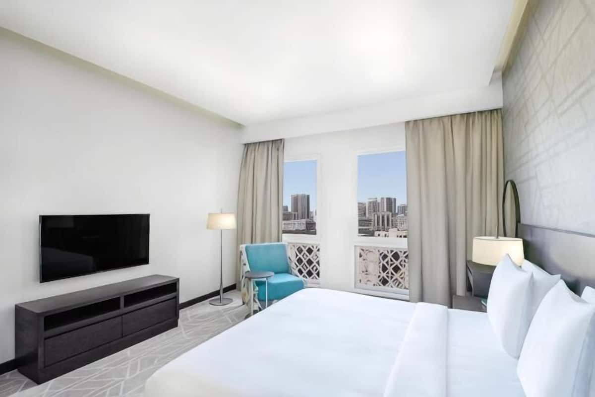 A hotel room with a large bed, a TV, a desk, and a balcony with a city view.