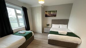 A hotel room with two single beds, a large window with curtains, a painting on the wall, and a bedside table with a lamp.