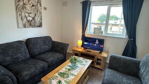A living room with a dark grey sofa set, a wooden coffee table with a tropical leaf pattern, a television on a wooden stand, and a painting of a lion on the wall.