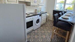 A compact kitchen with white cabinets, a washing machine, a dining area with a table and chairs, and a patterned floor.