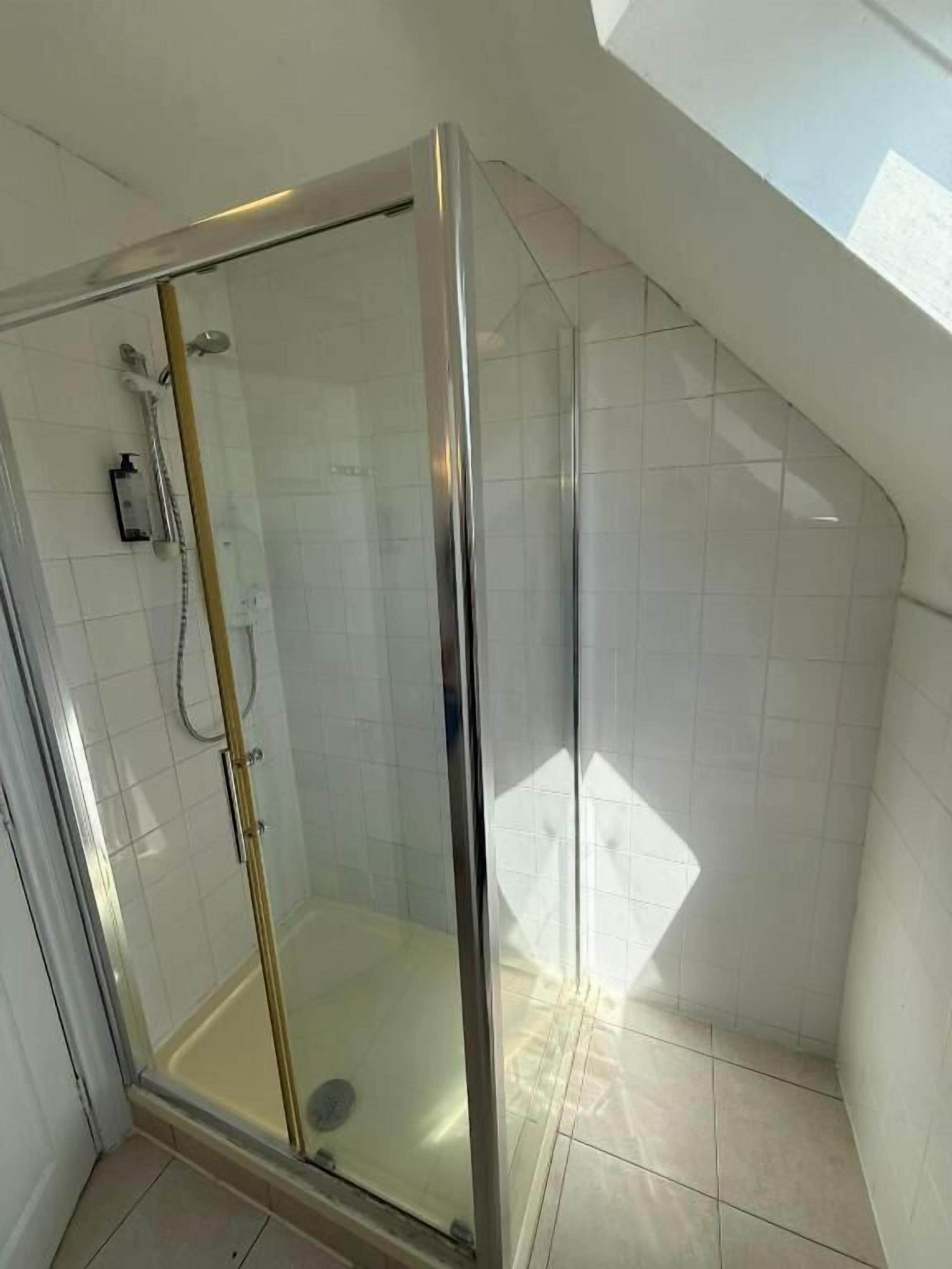 A corner shower with a glass door, white tiled walls, and a sloped ceiling.