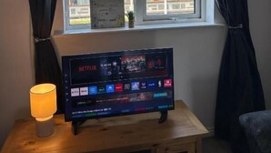 A room with a TV on a wooden table, a window with curtains, and a view of buildings outside.