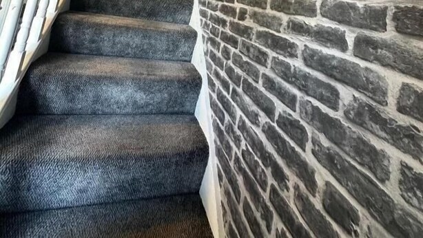 A staircase with a black carpet and white balusters, leading to a room with a brick wall and a painting on the wall.