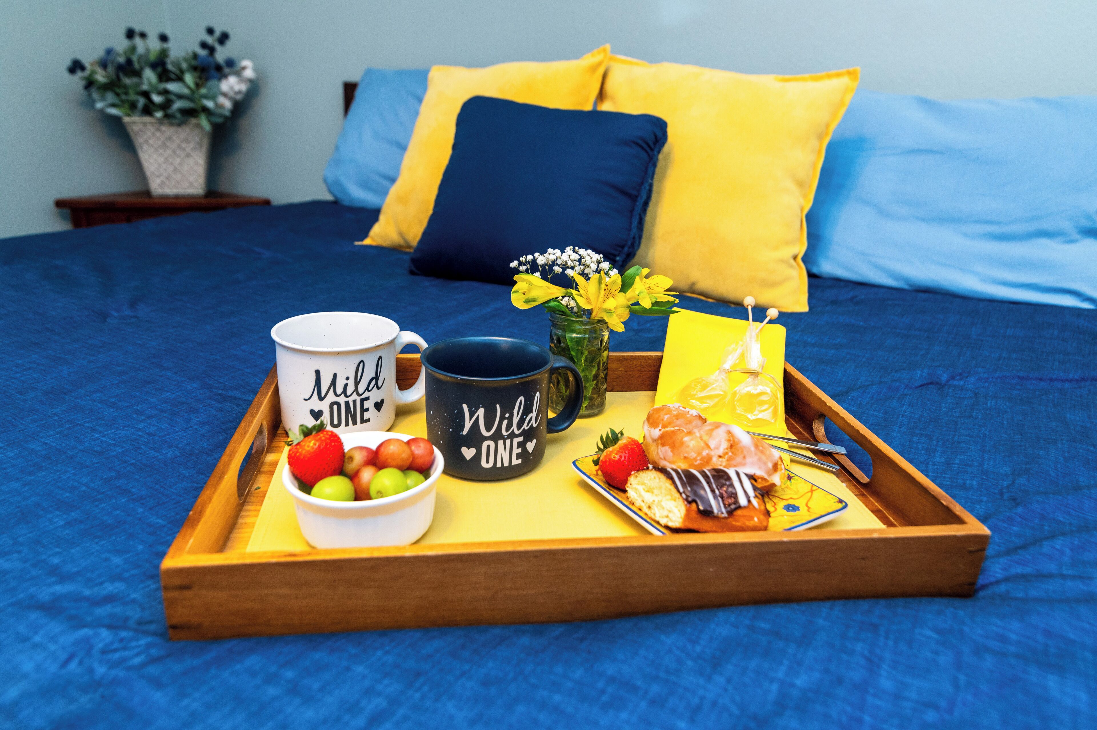 Cama com roupa de cama azul, bandeja com itens de café da manhã incluindo uma caneca, frutas e um pastel.