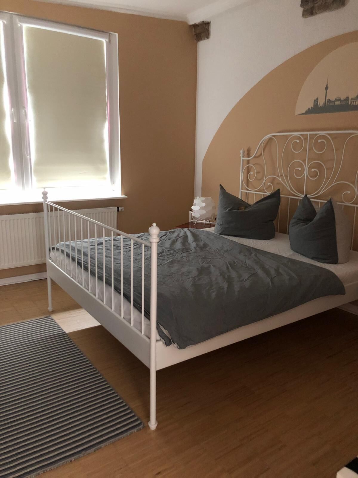 Une chambre avec un lit en métal blanc, une literie grise, une petite table de chevet avec une lampe, et une fresque représentant un paysage urbain sur le mur.