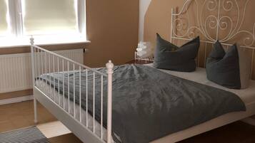 Une chambre avec un lit en métal blanc, une literie grise, une petite table de chevet avec une lampe, et une fresque représentant un paysage urbain sur le mur.