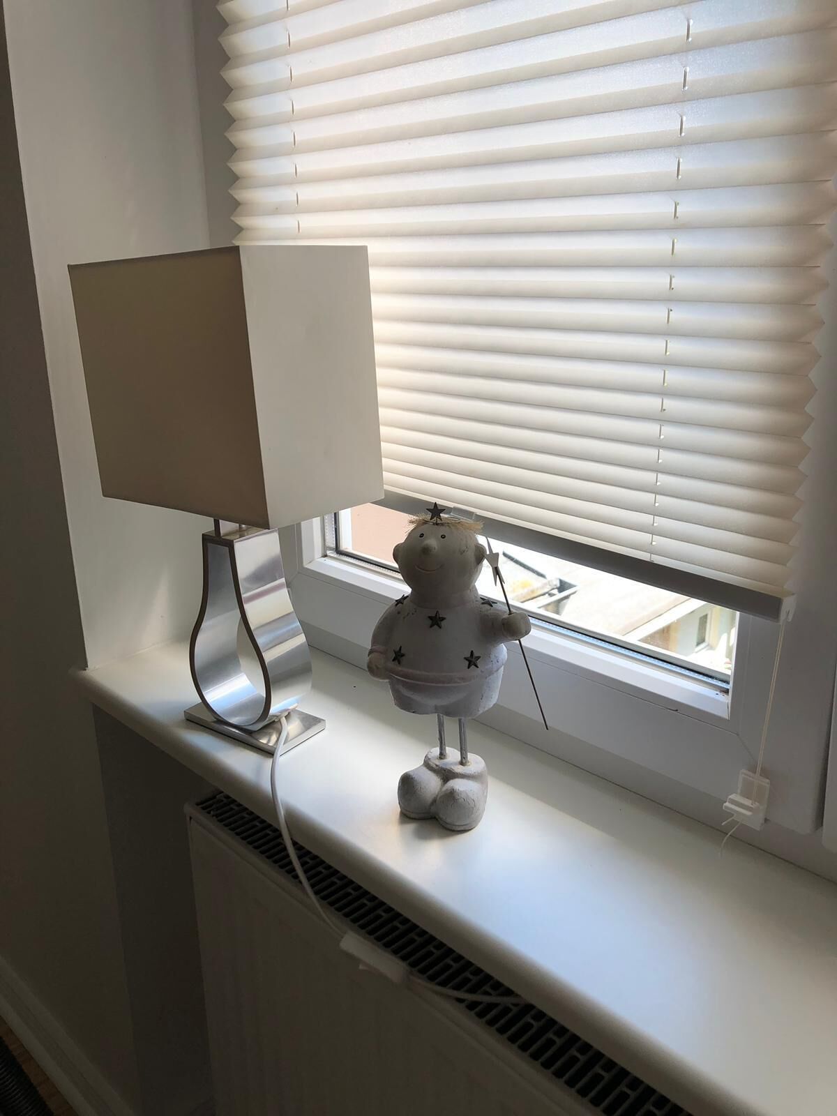 Ein Fenster mit weißen Jalousien, eine Lampe auf einem Tisch und eine kleine Figur auf dem Fensterbrett.