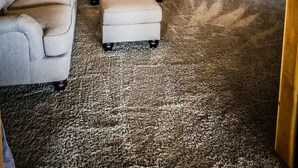 Una habitación con alfombra gris, un sofá gris, un sillón color beige, una pequeña mesita de madera y un techo con vigas de madera a la vista.