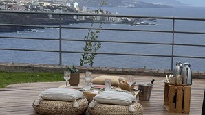 Une terrasse avec une table dressée pour le dîner, offrant une vue sur une ville côtière et l’océan.