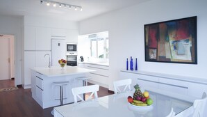 Une cuisine moderne avec des meubles blancs, un îlot central doté de tabourets, une table à manger avec des chaises et un panier de fruits.