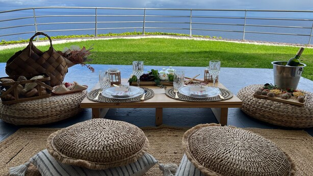 Un espace de restauration en plein air avec vue sur la mer, agrémenté de paniers en osier, d’une table en bois et de verres transparents.