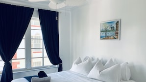 Ein ordentlich bezogenes Bett mit weißen Laken und einer dunkelblauen Decke, ein Deckenventilator und ein Bild einer Brücke und Gebäude an der Wand.