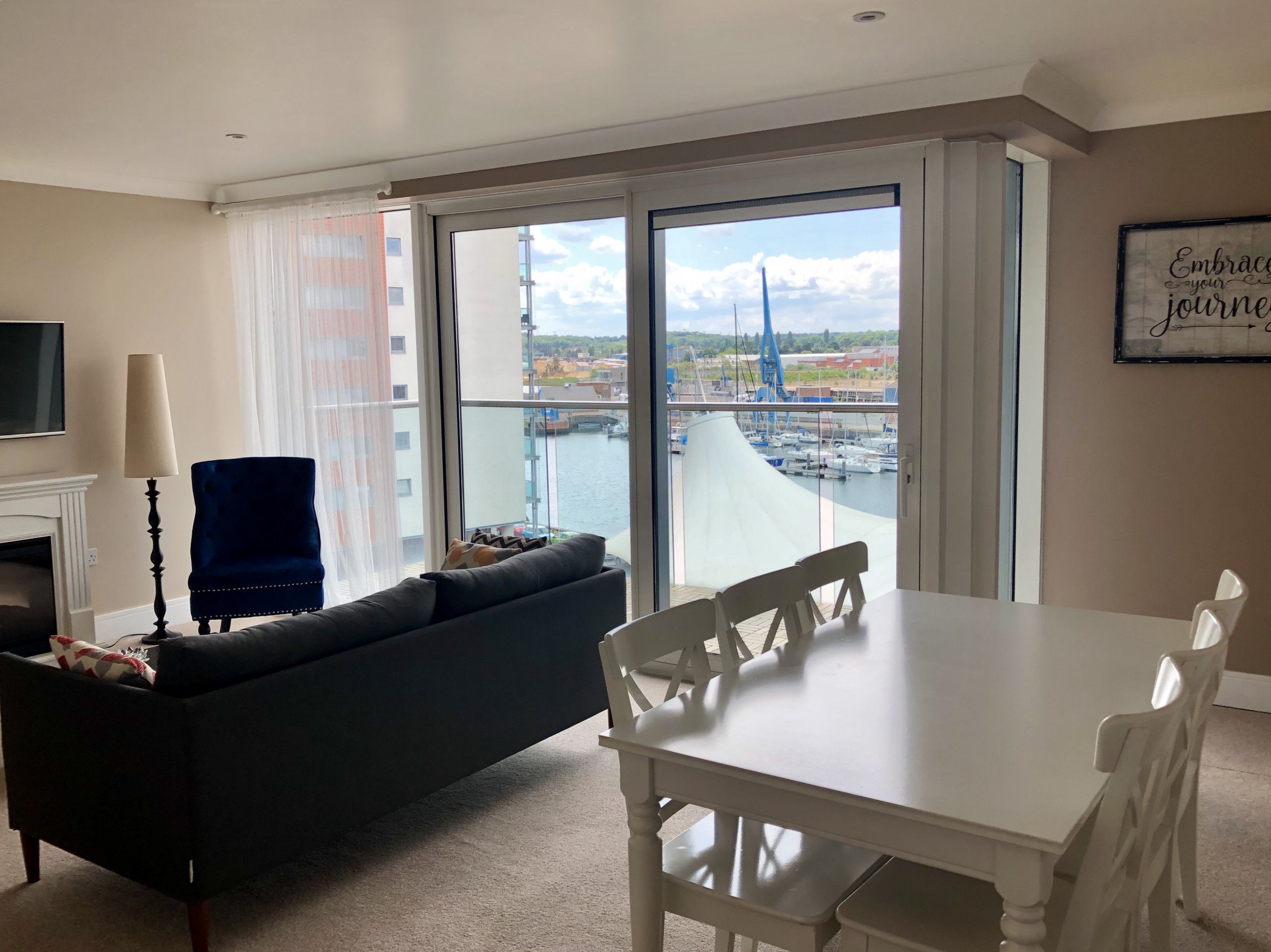A modern living room with a sofa, a dining table, and chairs. A large window offers a view of a marina and buildings.
