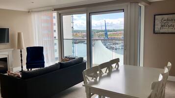 A modern living room with a sofa, a dining table, and chairs. A large window offers a view of a marina and buildings.