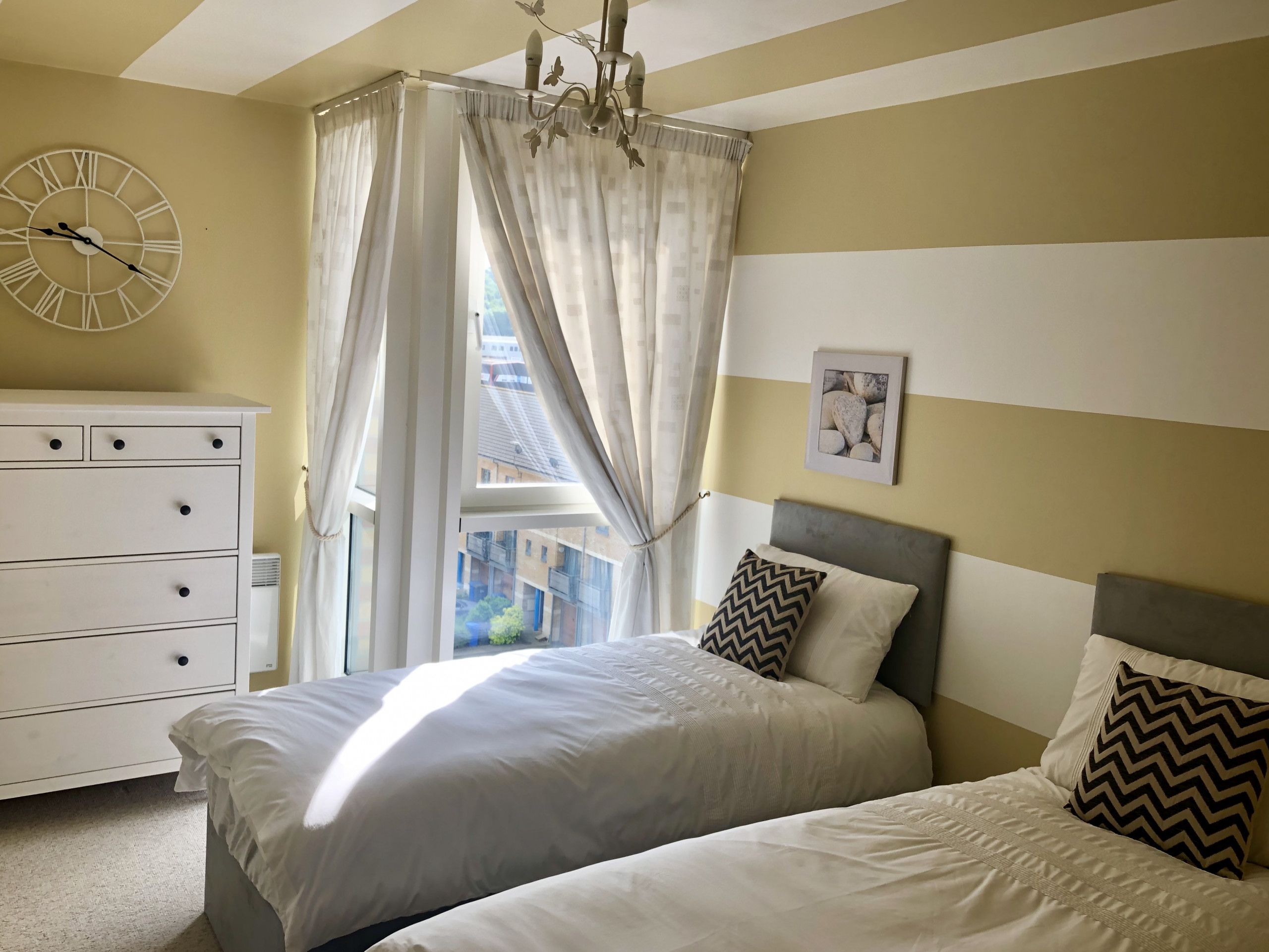 A bedroom with a bed, a dresser, a clock, and a window with curtains.