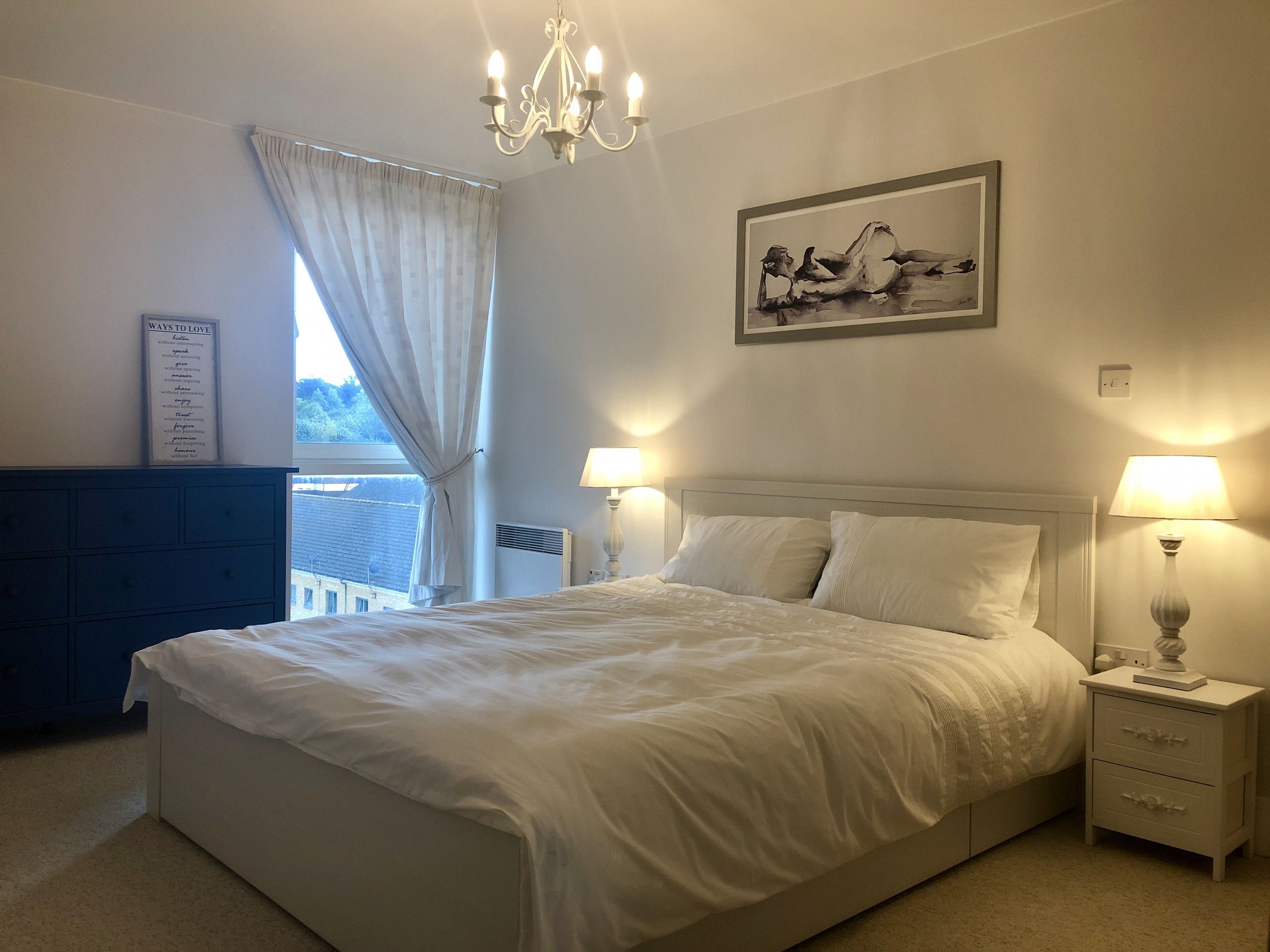 A bedroom with a large bed, two bedside lamps, a chandelier, a framed artwork, and a window with curtains.