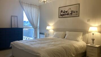A bedroom with a large bed, two bedside lamps, a chandelier, a framed artwork, and a window with curtains.