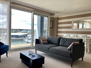 A modern living room with a grey sofa, a black coffee table, and a view of a marina through large windows. - Toothbrush Apartments - Ipswich Waterfront - Anchor St (Ipswich)