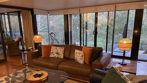 A modern living room with a leather sofa, a glass coffee table, and a view of trees through large windows. - Centrally located 2-bedroom luxury villa in Australia’s spa country (Hepburn Springs)