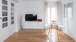 Una sala de estar con un televisor de pantalla plana, sillas blancas, una mesita con un frutero y una ventana con cortinas.