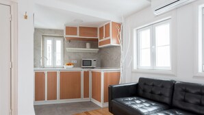 Una cocina compacta con armarios de madera, un microondas y un tazón de fruta sobre la encimera. Un sofá de cuero negro está ubicado cerca de una ventana.