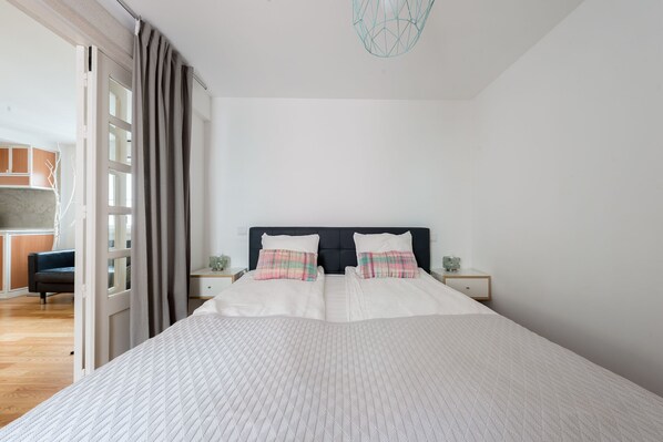 A modern bedroom with a large bed, two bedside tables, a dark headboard, and a view of a kitchen area through an open door.