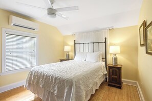 A bedroom with a bed, two bedside tables with lamps, a ceiling fan, an air conditioning unit, a window with blinds, and a wall-mounted picture frame.