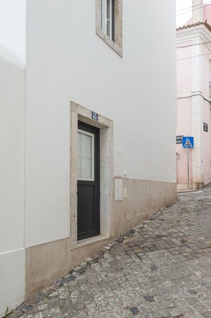 A narrow cobblestone street with a white building featuring a black door and a small window. - 0 Ophiussa (Lisbon)