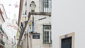 A narrow street with cobblestone pavement and a tram track. Buildings line the street, and there are signs and a flag visible. - 0 Ophiussa (Lisbon)
