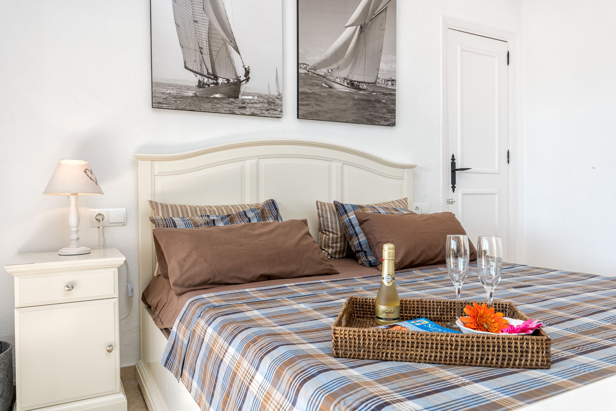 Cama bem arrumada com colcha listrada, mesa de cabeceira com abajur e bandeja com garrafa e copos na cama.