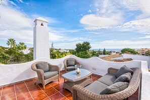 A rooftop terrace with wicker furniture, a glass table, and a view of the cityscape and sea.