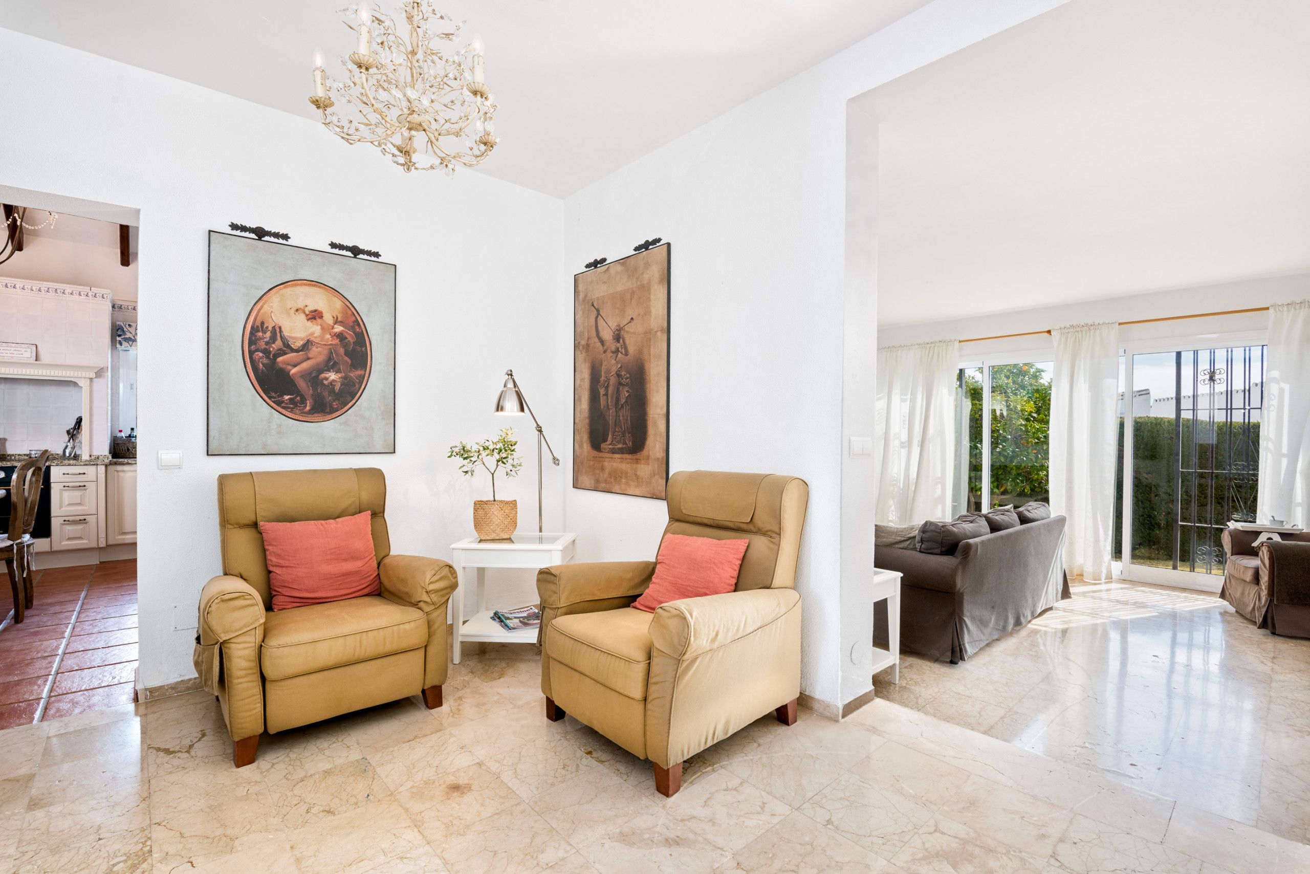 Un salon spacieux avec deux fauteuils beiges, une table d’appoint blanche et un canapé gris. Deux grandes œuvres d’art encadrées ornent les murs, ainsi qu’un lustre. De grandes fenêtres offrent une vue dégagée.