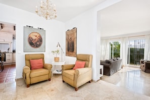 A spacious living room with two beige armchairs, a white side table, and a grey sofa. There are two large framed artworks on the walls, a chandelier, and a view through large windows.