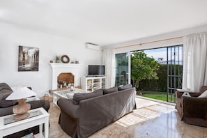 A modern living room with a grey sofa, a white coffee table, and a flat-screen TV. There is a fireplace with decorative items and a large window with curtains.